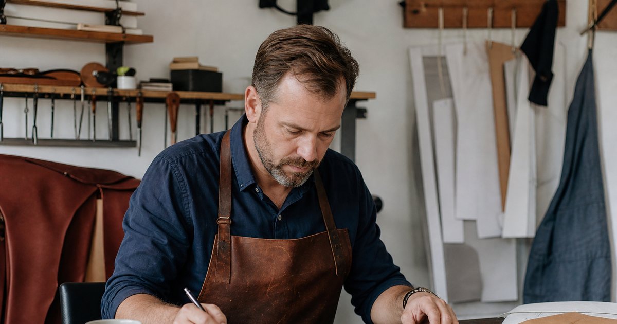 A leatherworker in a small workshop sitting at a desk with a closed notebook, a coffee mug and a stack of envelopes, tools and hides hanging on the wall behind